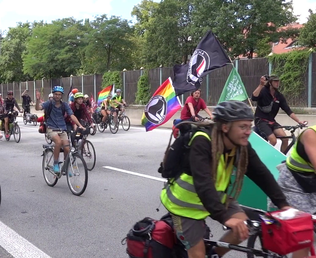 Auf Autobahn-Abschnitt fahren OKNB-Teilnehmende an Kamera vorbei, drei mit großen Antifa-Flaggen.